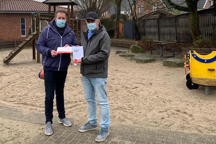 Zwei Personen mit Mund-Nasen-Schutz stehen auf einem Sandspielplatz und halten ein Dokument in der Hand.