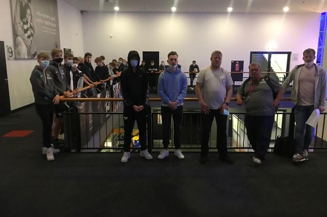 Gruppenszene von jungen Personen und Erwachsenen in einem Innenbereich, alle tragen Masken und stehen an einem Gel&auml;nder.