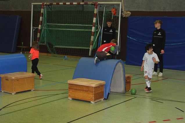 Kinder spielen in einer Sporthalle, w&auml;hrend zwei in Trainingsanz&uuml;gen zuschauen. Eine Person springt &uuml;ber ein Hindernis.