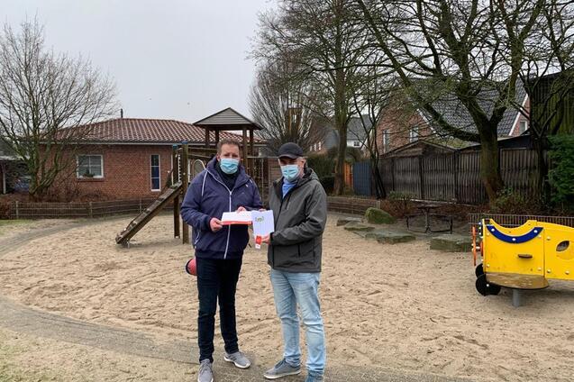 Zwei Personen mit Mund-Nasen-Schutz stehen auf einem Sandspielplatz und halten ein Dokument in der Hand.