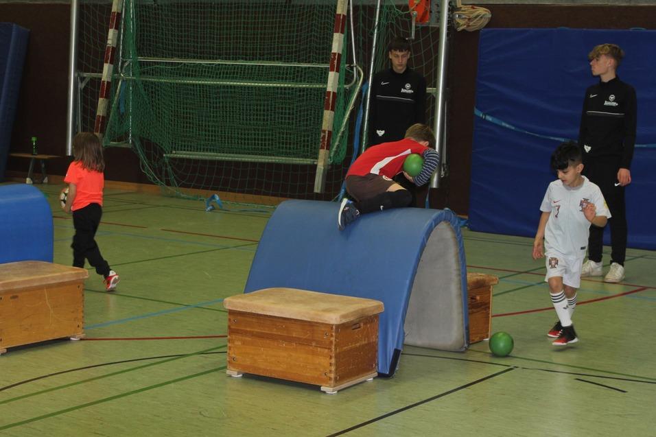 Kinder spielen in einer Sporthalle, w&auml;hrend zwei in Trainingsanz&uuml;gen zuschauen. Eine Person springt &uuml;ber ein Hindernis.