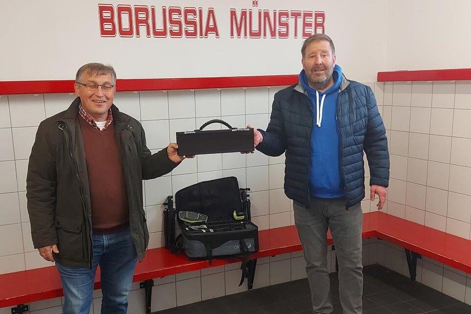 Zwei M&auml;nner stehen in einem Umkleideraum von Borussia M&uuml;nster, einer h&auml;lt eine schwarze Tasche, der andere tr&auml;gt einen blauen Hoodie.
