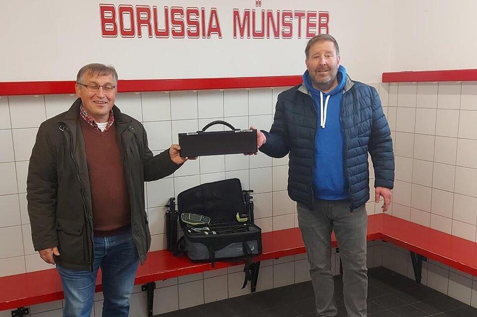 Zwei M&auml;nner stehen in einem Umkleideraum von Borussia M&uuml;nster, einer h&auml;lt eine schwarze Tasche, der andere tr&auml;gt einen blauen Hoodie.