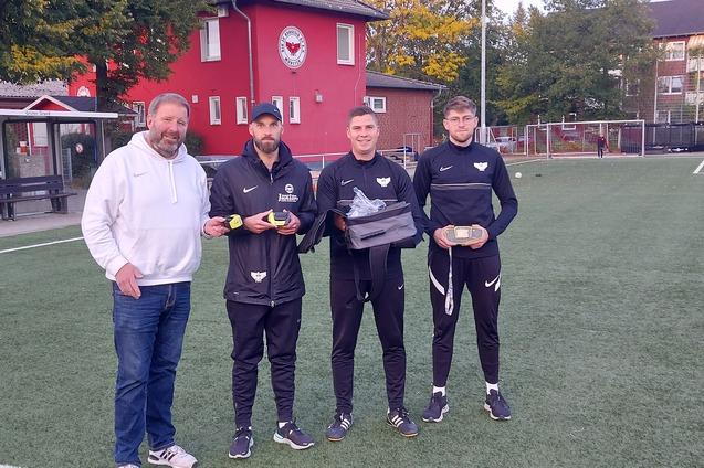 Vier Personen stehen auf einem Fu&szlig;ballplatz; zwei Spieler halten Preise, w&auml;hrend ein Trainer daneben steht.