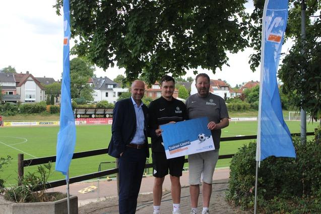 Drei M&auml;nner stehen auf einem Sportgel&auml;nde, halten ein Plakat und posieren vor einem Fu&szlig;ballplatz mit B&auml;umen im Hintergrund.