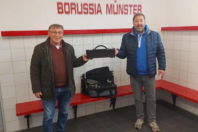 Zwei M&auml;nner stehen in einem Umkleideraum von Borussia M&uuml;nster, einer h&auml;lt eine schwarze Tasche, der andere tr&auml;gt einen blauen Hoodie.