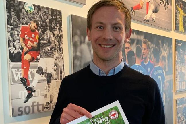 Mann steht vor einer Wand mit Fu&szlig;ballfotos und h&auml;lt ein Plakat der Borussia & M&uuml;nster Juniors in der Hand.