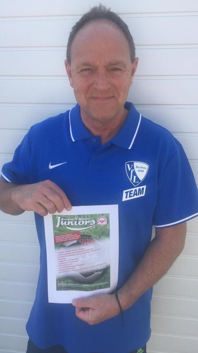 Mann in blauem Team-Trikot h&auml;lt ein Informationsblatt &uuml;ber eine Juniorenveranstaltung vor einer hellen Wand.