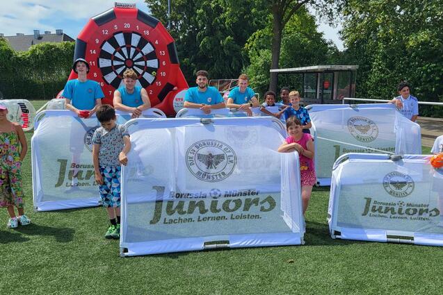 Gruppe von Kindern und Betreuern auf einem Sportplatz hinter transparenten Spielschildern mit der Aufschrift &bdquo;Juniors&ldquo;.