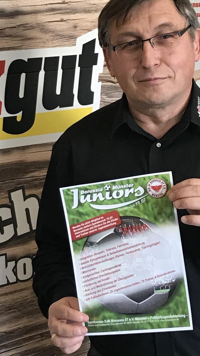 Mann h&auml;lt ein Informationsblatt &uuml;ber die Jugendabteilung von Borussia M&ouml;nster vor einer Werbewand.