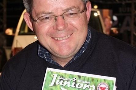 Mann mit Brille l&auml;chelt und h&auml;lt eine Zeitschrift mit dem Titel &bdquo;Borussia M&uuml;nster Juniors&ldquo; in der Hand.