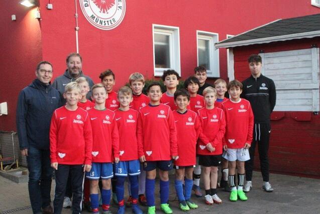 Gruppe von zehn Jugendlichen in roten Trikots, posierend vor einer roten Wand mit Trainern und Betreuern im Hintergrund.