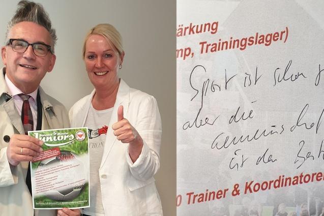 Zwei Personen posieren l&auml;chelnd mit einem Trainingslager-Flyer, daneben ein handschriftlicher Text auf Papier.