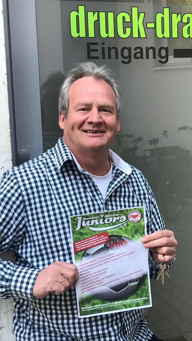 Mann mit grauem Haar h&auml;lt ein Plakat vor einem Eingang mit der Aufschrift &bdquo;druck-drauf&ldquo;. Fahrr&auml;der sind im Hintergrund sichtbar.