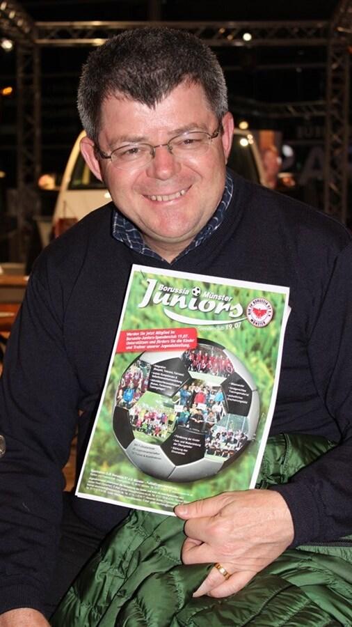 Mann mit Brille h&auml;lt eine Brosch&uuml;re von Borussia M&uuml;nster Juniors in der Hand, l&auml;chelnd vor einer Holzplatte.