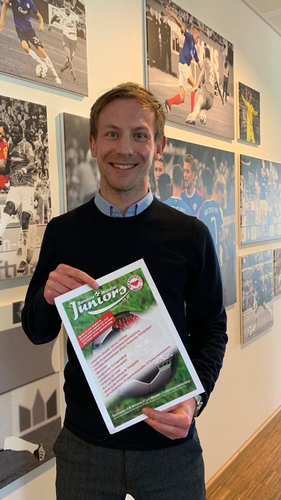 Mann steht vor einer Wand mit Fu&szlig;ballfotos und h&auml;lt ein Plakat der Borussia & M&uuml;nster Juniors in der Hand.