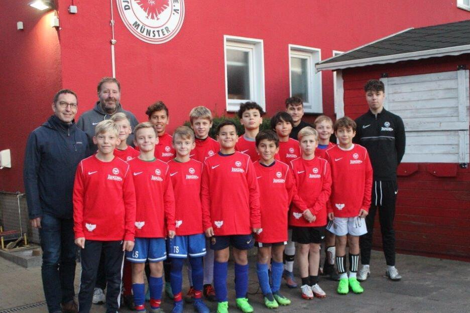 Gruppe von zehn Jugendlichen in roten Trikots, posierend vor einer roten Wand mit Trainern und Betreuern im Hintergrund.