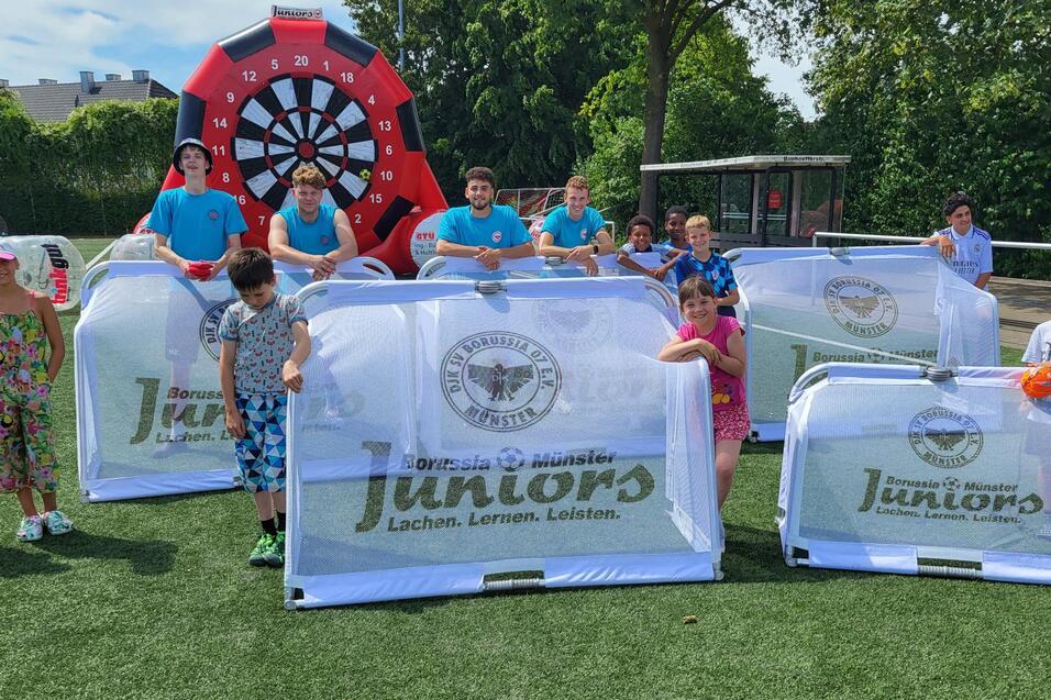 Gruppe von Kindern und Betreuern auf einem Sportplatz hinter transparenten Spielschildern mit der Aufschrift &bdquo;Juniors&ldquo;.