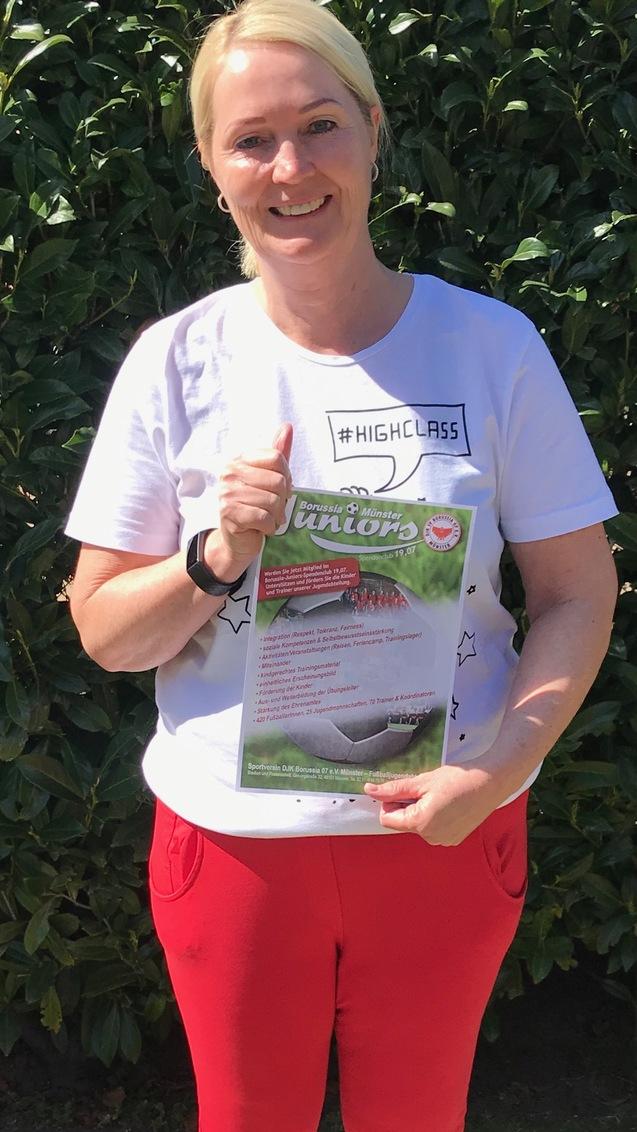Frau in wei&szlig;em T-Shirt und roten Hosen h&auml;lt ein Plakat mit Informationen, vor einer Hecke im Freien.