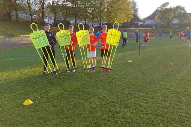 F&uuml;nf Kinder in Sportkleidung halten leuchtend gelbe Trainingspuppen auf einem Fu&szlig;ballplatz bei Sonnenuntergang.