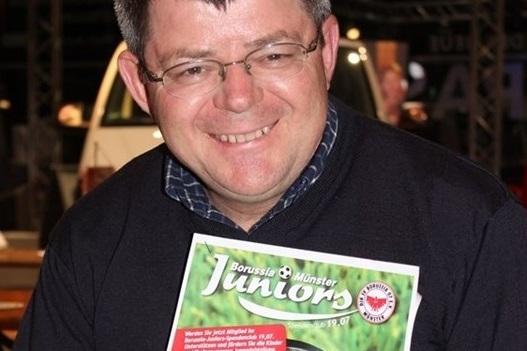 Mann mit Brille l&auml;chelt und h&auml;lt eine Zeitschrift mit dem Titel &bdquo;Borussia M&uuml;nster Juniors&ldquo; in der Hand.