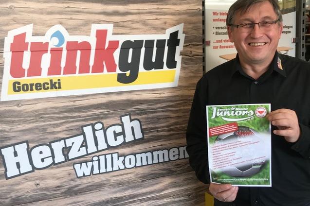 Mann h&auml;lt ein Informationsblatt vor einem plakatierten Hintergrund mit der Aufschrift &bdquo;trinkgut Gorecki&ldquo; und &bdquo;Herzlich willkommen&ldquo;.