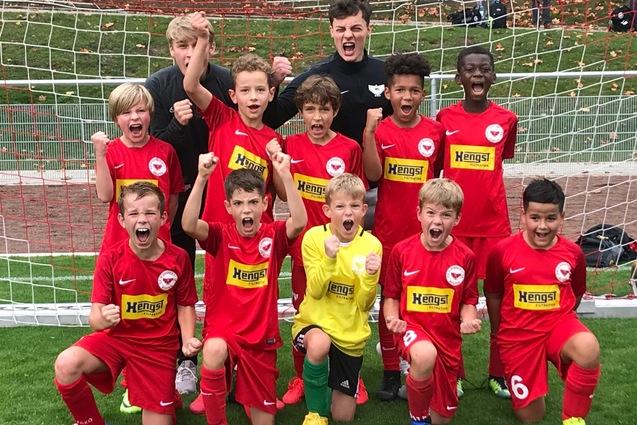 Gruppe jubelnder junger Fu&szlig;ballspieler in roten Trikots posiert vor einem Fu&szlig;balltor auf einem gr&uuml;nen Rasenplatz.