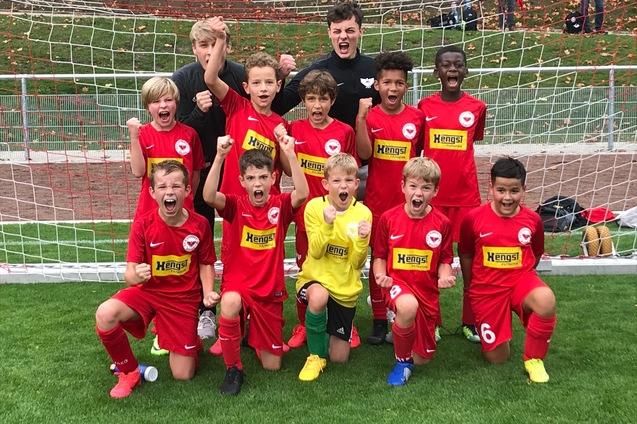 Gruppe jubelnder junger Fu&szlig;ballspieler in roten Trikots posiert vor einem Fu&szlig;balltor auf einem gr&uuml;nen Rasenplatz.