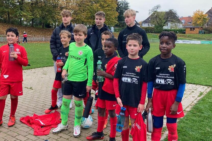 Gruppe von zehn Kindern in Fu&szlig;balltrikots, die auf einem Platz im Freien posieren, mit B&auml;umen im Hintergrund.