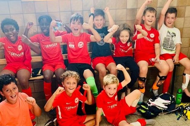 Gruppe junger Fu&szlig;ballspieler in roten Trikots sitzt fr&ouml;hlich auf einer Bank und jubelt in einem Umkleideraum.