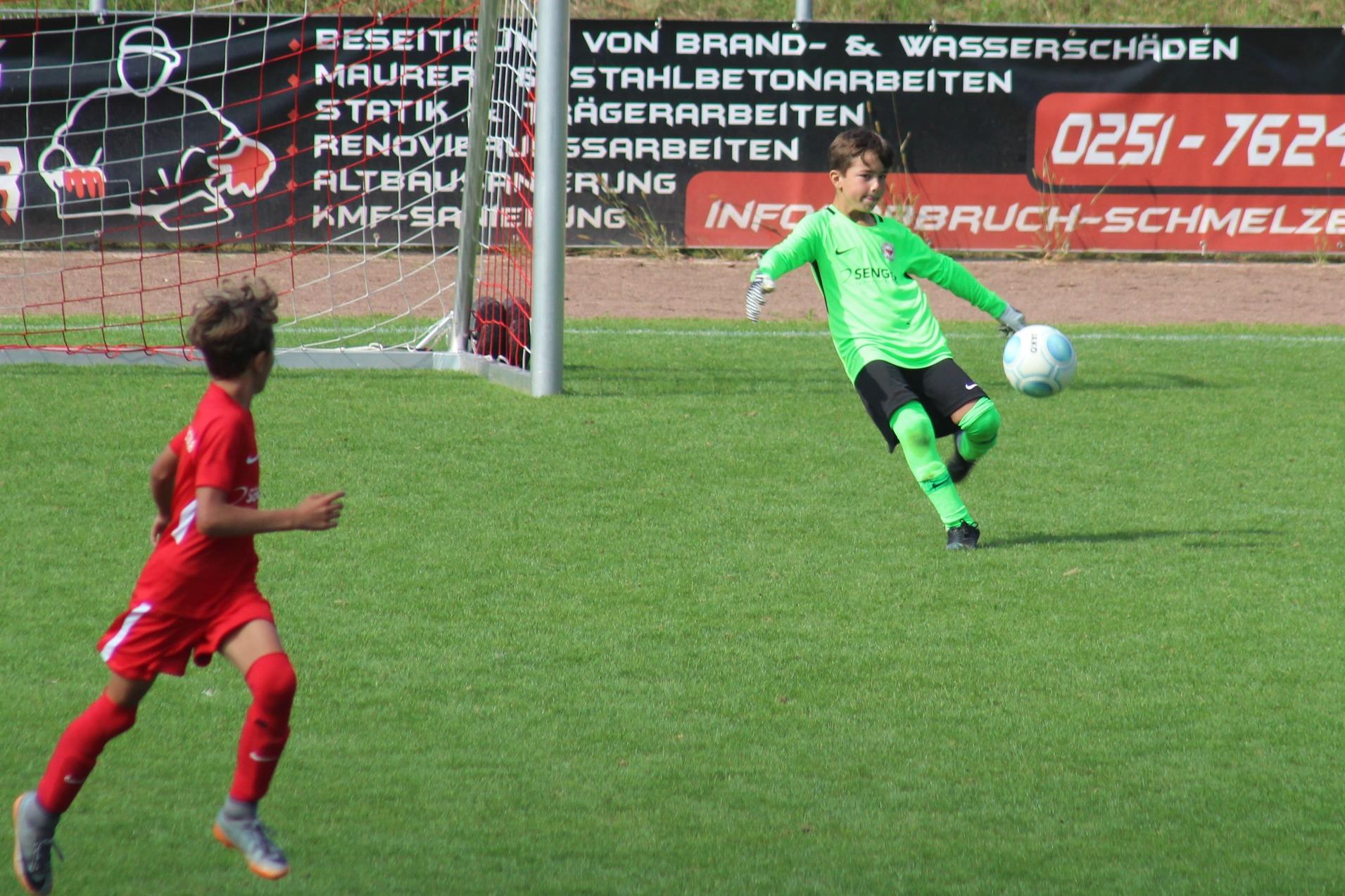 Zwei Kinder im Fu&szlig;ballspiel, ein Torwart in Gr&uuml;n f&auml;ngt einen Ball, w&auml;hrend ein Spieler in Wei&szlig; darauf zielt.