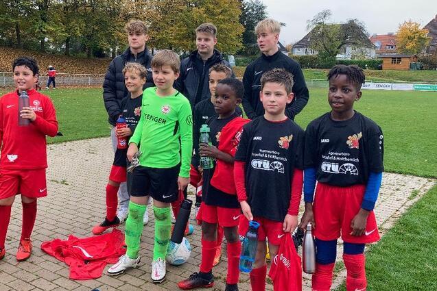 Gruppe von zehn Kindern in Fu&szlig;balltrikots, die auf einem Platz im Freien posieren, mit B&auml;umen im Hintergrund.
