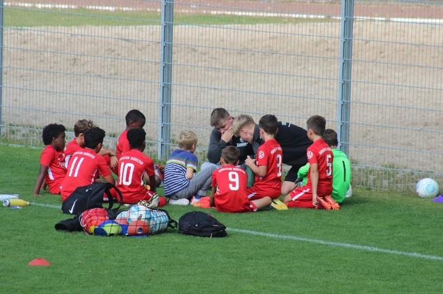 Gruppe von Kindern in roten Fu&szlig;balltrikots sitzt auf dem Rasen, um einen Trainer, der ihnen Anweisungen gibt.
