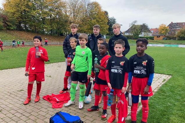 Gruppe von zehn Kindern in Fu&szlig;balltrikots, die auf einem Platz im Freien posieren, mit B&auml;umen im Hintergrund.