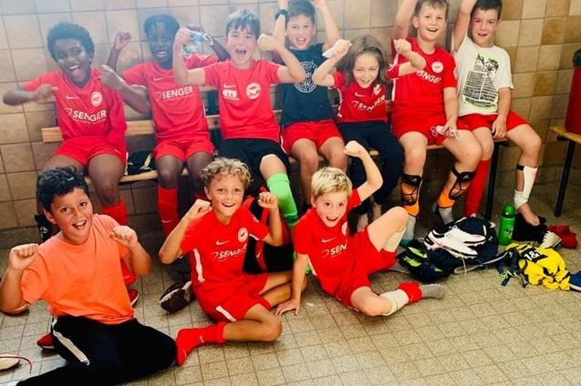 Gruppe junger Fu&szlig;ballspieler in roten Trikots sitzt fr&ouml;hlich auf einer Bank und jubelt in einem Umkleideraum.