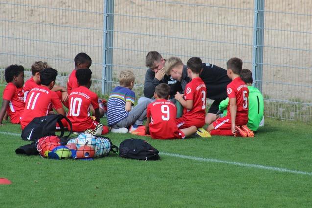 Gruppe von Kindern in roten Fu&szlig;balltrikots sitzt auf dem Rasen, um einen Trainer, der ihnen Anweisungen gibt.