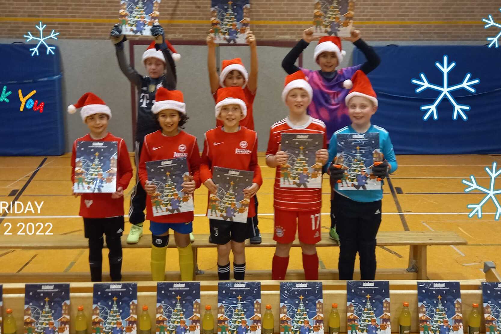 Gruppe von acht Kindern in Weihnachtsm&uuml;tzen, die stolz ihre Plakate auf einer Bank in einer Sporthalle zeigen.