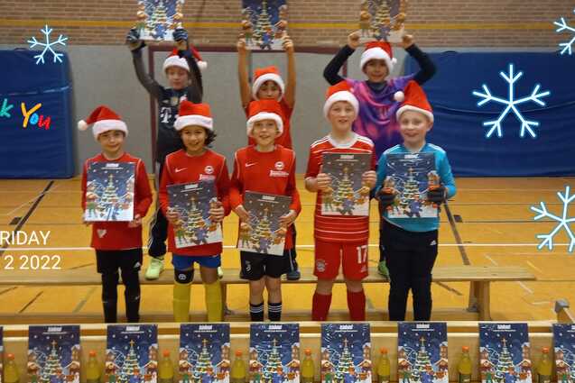 Gruppe von acht Kindern in Weihnachtsm&uuml;tzen, die stolz ihre Plakate auf einer Bank in einer Sporthalle zeigen.