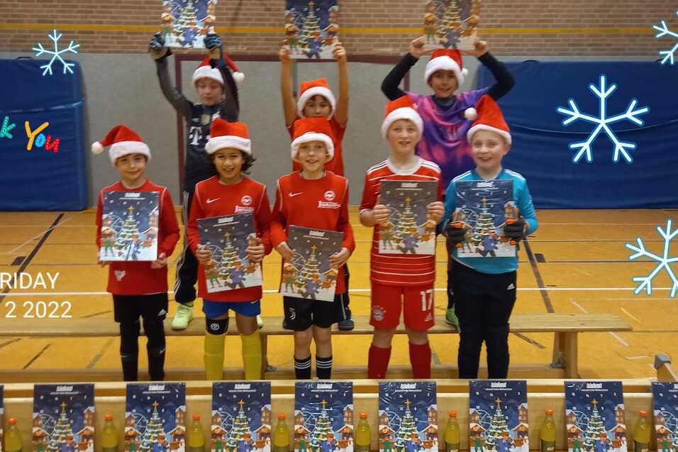 Gruppe von acht Kindern in Weihnachtsm&uuml;tzen, die stolz ihre Plakate auf einer Bank in einer Sporthalle zeigen.