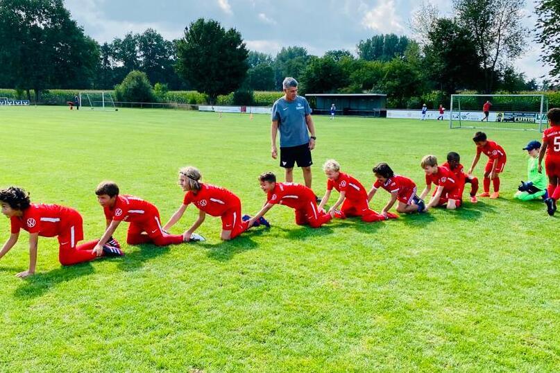 Gruppe von Kindern in roten Trikots, die auf einem Fu&szlig;ballplatz in einer Reihe krabbeln, betreut von einem Trainer.