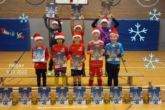 Gruppe von acht Kindern in Weihnachtsm&uuml;tzen, die stolz ihre Plakate auf einer Bank in einer Sporthalle zeigen.