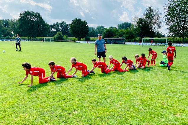 Gruppe von Kindern in roten Trikots, die auf einem Fu&szlig;ballplatz in einer Reihe krabbeln, betreut von einem Trainer.
