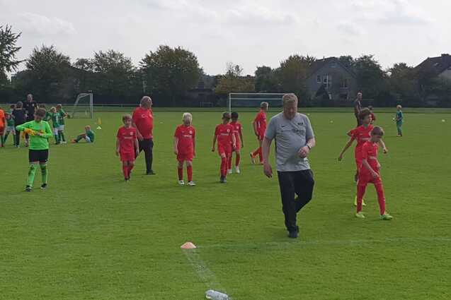 Fu&szlig;ballmannschaft in roten Trikots joggt auf einem gr&uuml;nen Spielfeld, w&auml;hrend Trainer sie anleitet.