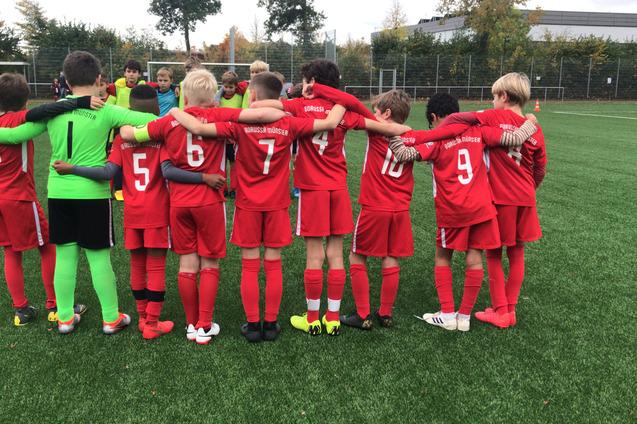 Eine Gruppe junger Fu&szlig;ballspieler in roten Trikots steht mit dem R&uuml;cken zueinander und umarmt sich auf einem Kunstrasenplatz.
