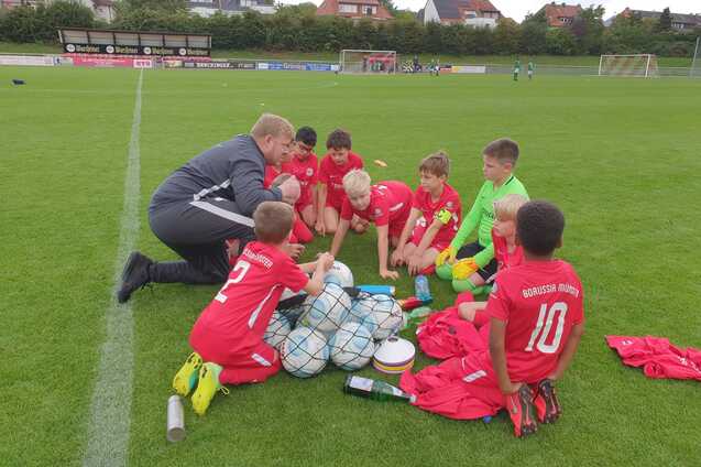 Gruppe von Kindern in roten Trikots sitzt um B&auml;lle und Ausr&uuml;stung auf einem Fu&szlig;ballfeld, w&auml;hrend ein Trainer spricht.