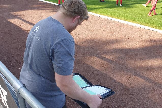 Ein Trainer beobachtet ein Fu&szlig;ballspiel und notiert auf einem Tablet, w&auml;hrend Spieler auf dem Feld trainieren.