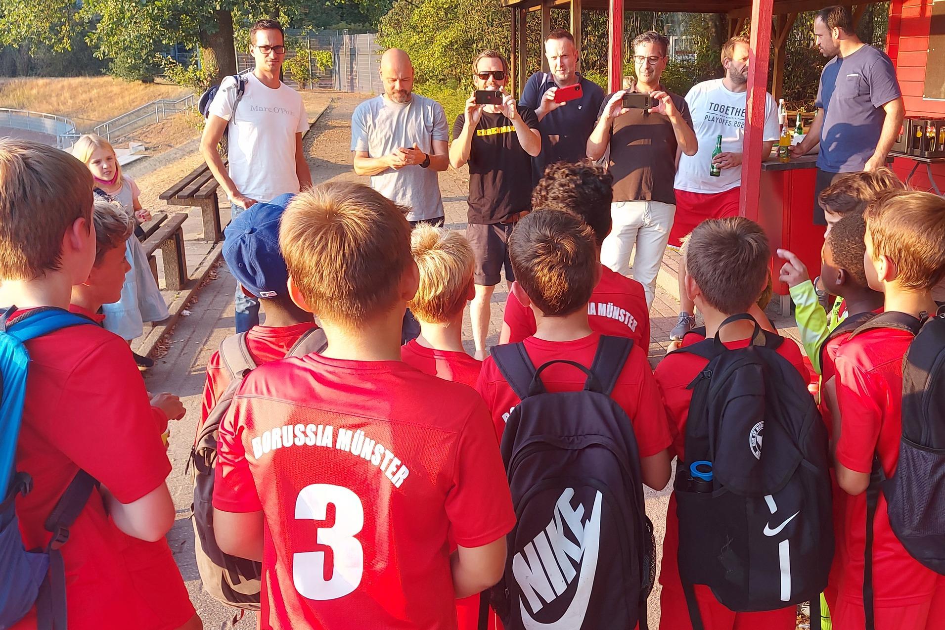 Gruppe von Kindern im roten Fu&szlig;balltrikot h&ouml;rt einem Trainer an einem Vereinsheim zu.