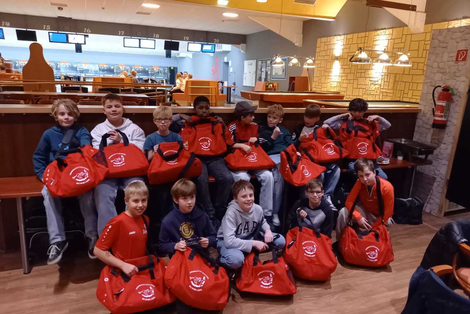 Gruppe von Jungen sitzt mit roten Sporttaschen in einem Bowling-Caf&eacute;, im Hintergrund sind Bowlingbahnen sichtbar.