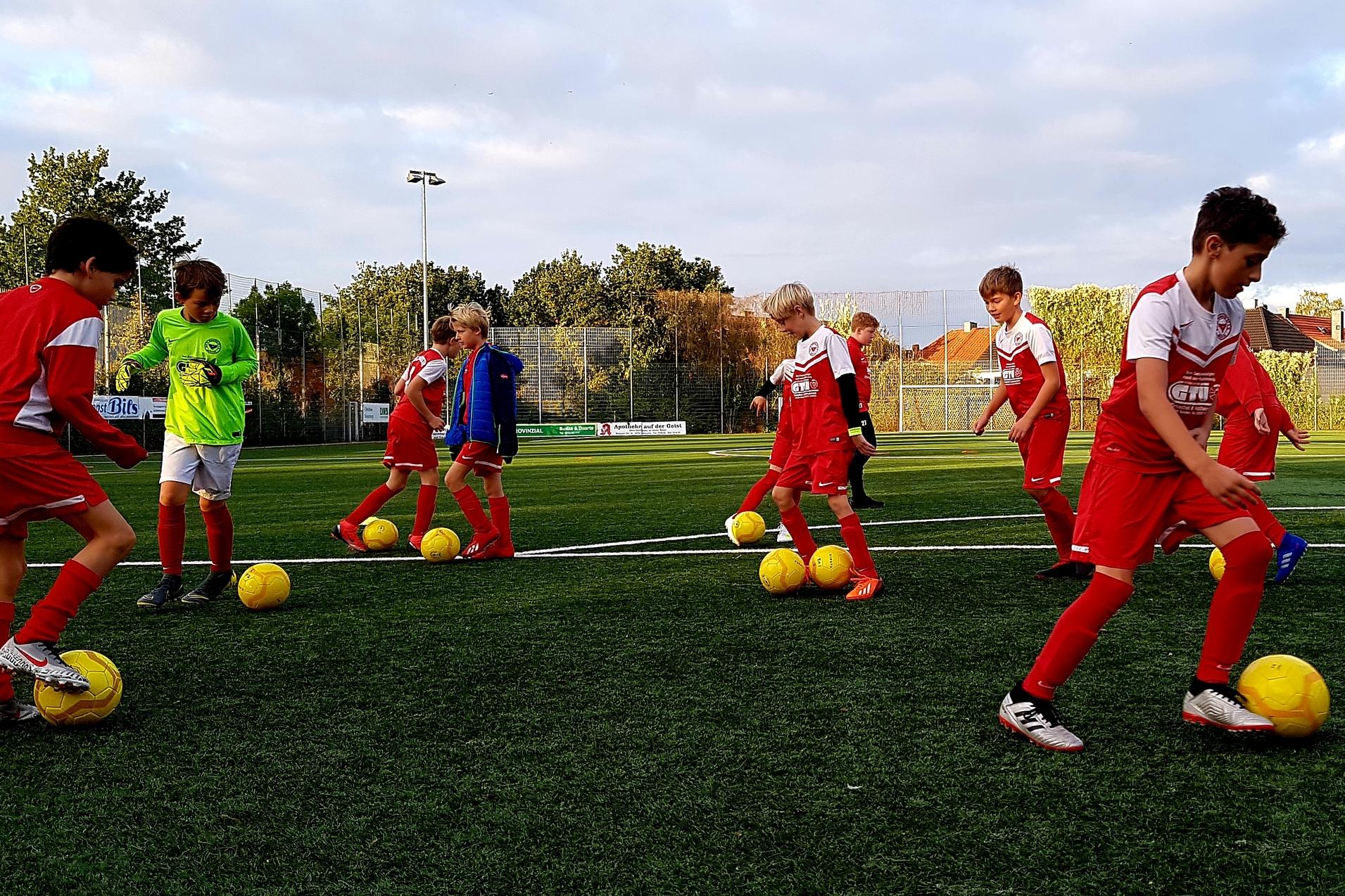 Jugendliche Fu&szlig;ballspieler in roten Trikots &uuml;ben mit gelben B&auml;llen auf einem Kunstrasenplatz unter bew&ouml;lktem Himmel.