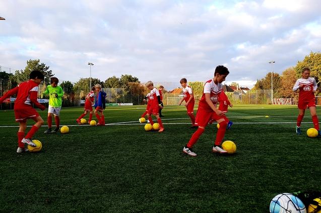 Jugendliche Fu&szlig;ballspieler in roten Trikots &uuml;ben mit gelben B&auml;llen auf einem Kunstrasenplatz unter bew&ouml;lktem Himmel.